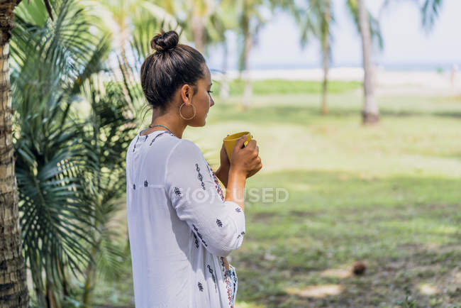 Vista laterale donna tranquilla che beve caffè dalla tazza e in piedi vicino alla palma sulla spiaggia soleggiata in Costa Rica — Foto stock