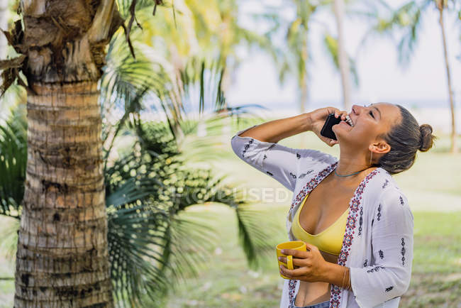Mulher feliz falando no smartphone olhando embora enquanto segurando caneca e de pé no gramado ensolarado por palmeira na Costa Rica — Fotografia de Stock