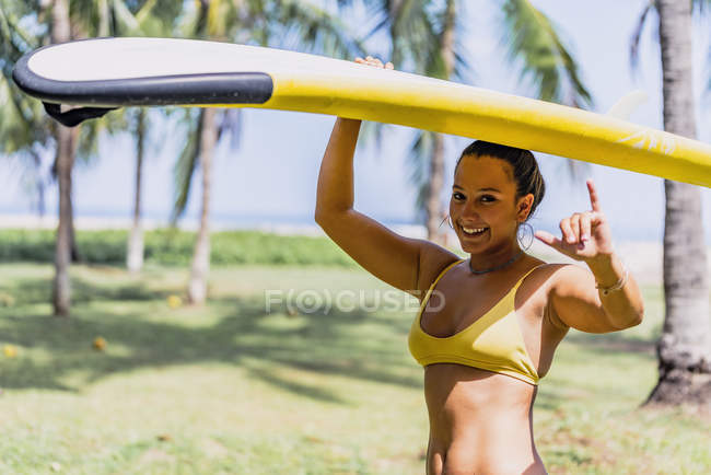 Positive fit woman in swimsuit carrying yellow paddleboard over the head in sunny seashore by palm trees in Costa Rica — Stock Photo