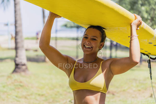 Mujer en traje de baño en forma positiva llevando paddleboard amarillo sobre la cabeza en la costa soleada por palmeras en Costa Rica - foto de stock
