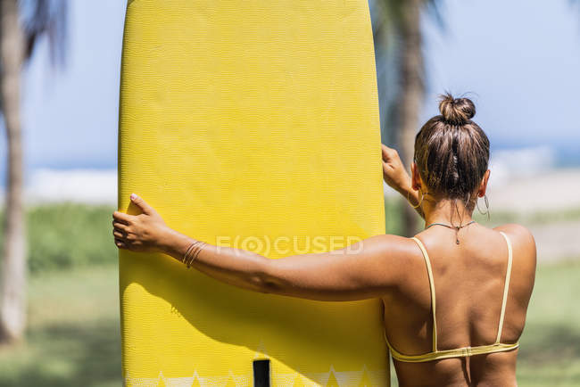 Back view of woman in swimsuit holding yellow paddleboard and standing in sunny seashore by palm trees in Costa Rica — Stock Photo