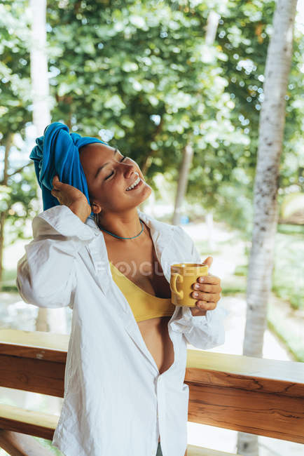 Contenuto donna in testa avvolgere godendo bevanda calda mentre in piedi da ringhiera in legno e guardando alberi frondosi in Costa Rica — Foto stock