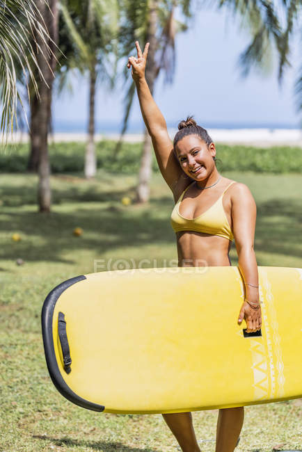 Positive fit woman in swimsuit holding yellow paddleboard and standing in sunny seashore by palm trees in Costa Rica — Stock Photo