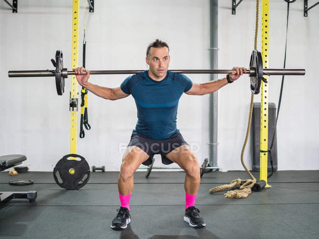 Приседающий спокойный спортсмен в спортивной одежде, тренирующийся со штангой и смотрящий в сторону белой стены — стоковое фото