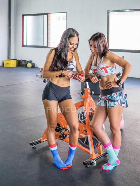 Весёлые спортсменки в спортивной одежде фокусируются и взаимодействуют со смартфоном, стоя на стационарном оранжевом велосипеде — стоковое фото
