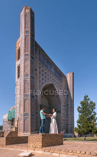 Vista lateral do homem e da mulher de mãos dadas enquanto estão do lado de fora Bibikhanum Hotel contra o céu azul sem nuvens em Samarcanda, Uzbequistão — Fotografia de Stock
