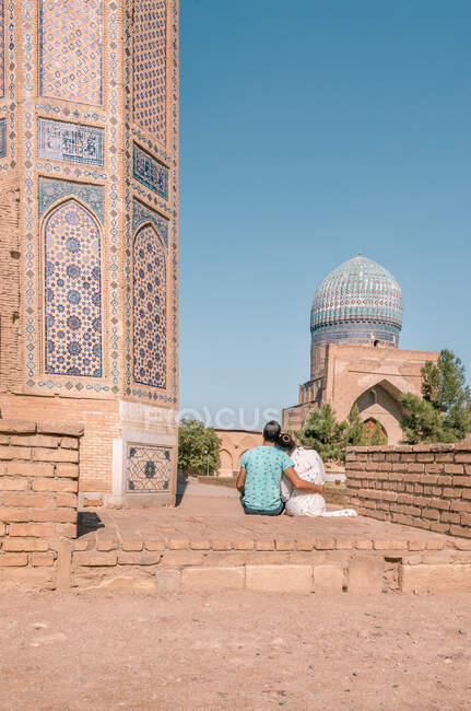 Visão traseira do casal abraçando uns aos outros enquanto se sentam fora do edifício tradicional contra o céu azul sem nuvens em Samarcanda, Uzbequistão — Fotografia de Stock