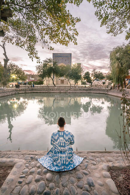 Visão traseira da fêmea em vestuário ornamental sentado perto de água tranquila da piscina em pacífico jardim à noite em Bukhara, Uzbequistão — Fotografia de Stock