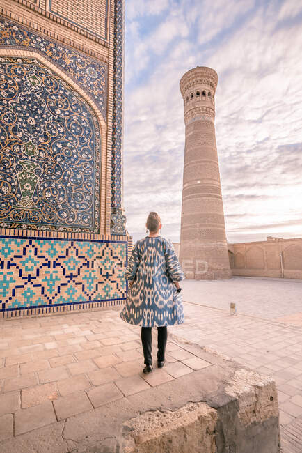 Vista posteriore della femmina in cappotto ornamentale in piedi sul quadrato invecchiato contro cielo nuvoloso a Bukhara, Uzbekistan — Foto stock