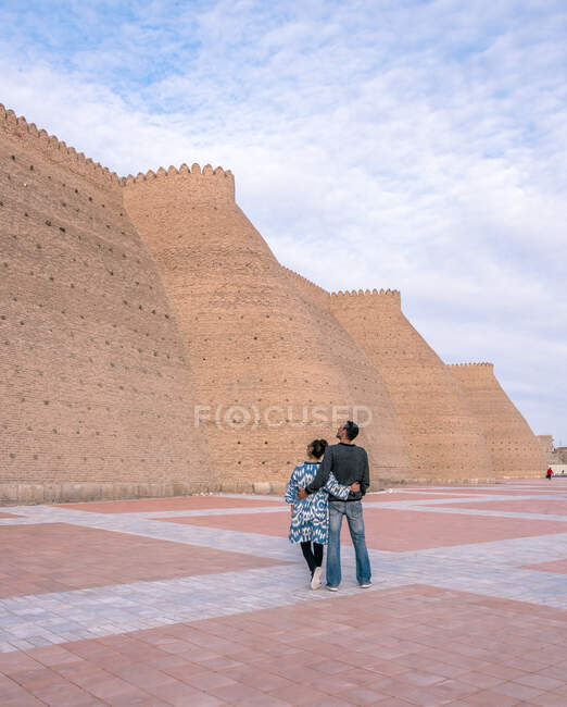 Visão traseira do homem étnico e da mulher abraçando um ao outro e olhando em paredes rasgadas da fortaleza antiga no dia nublado em Bukhara, Uzbequistão — Fotografia de Stock