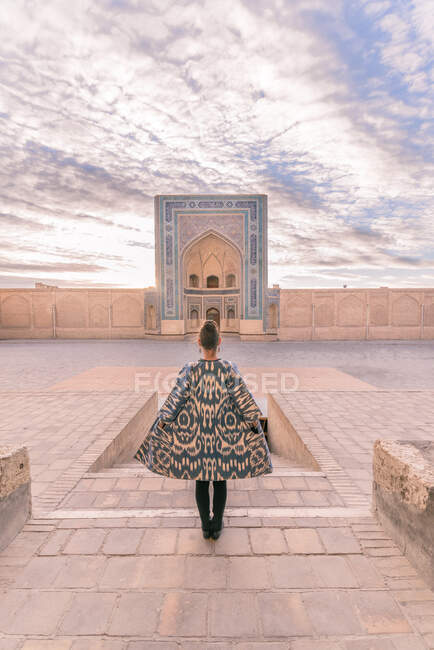 Visão traseira da fêmea em casaco ornamental em pé na praça envelhecida contra o céu nublado em Bucara, Uzbequistão — Fotografia de Stock
