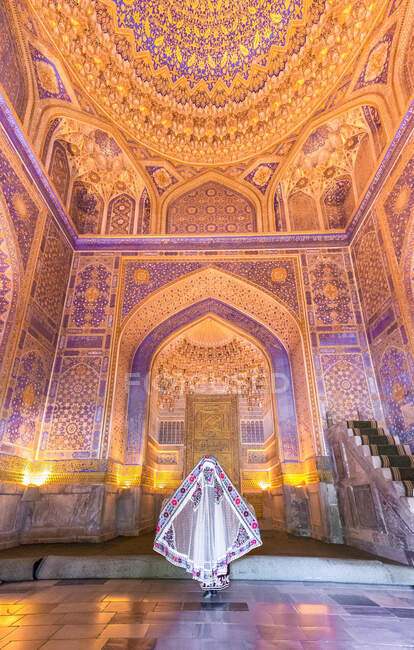 Rückansicht einer anonymen Frau in traditionellem Gewand, die inmitten eines beleuchteten ornamentalen Gebäudes steht, während sie Registan in Samarkand, Usbekistan besucht — Stockfoto