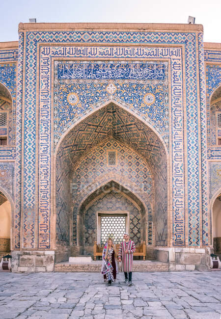 Paar in traditioneller Kleidung steht vor gewölbtem ornamentalem Gebäude bei einem Besuch des Registan in Samarkand, Usbekistan — Stockfoto