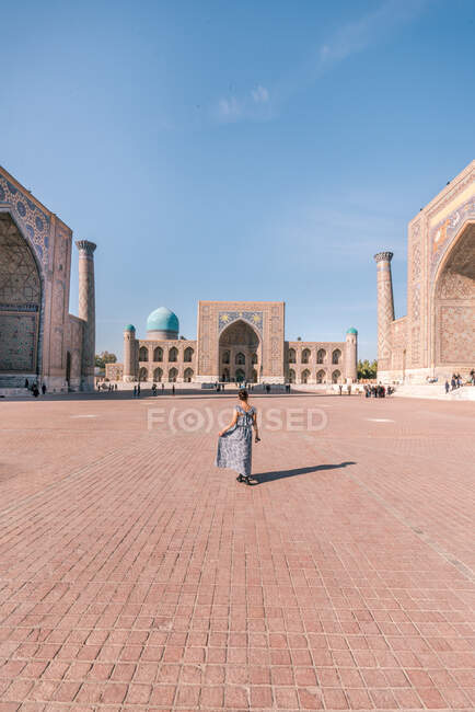 Vista posteriore di donna anonima in abito camminare sulla piazza lastricata vicino a edifici islamici tradizionali durante la visita Registan in Samarcanda, Uzbekistan — Foto stock