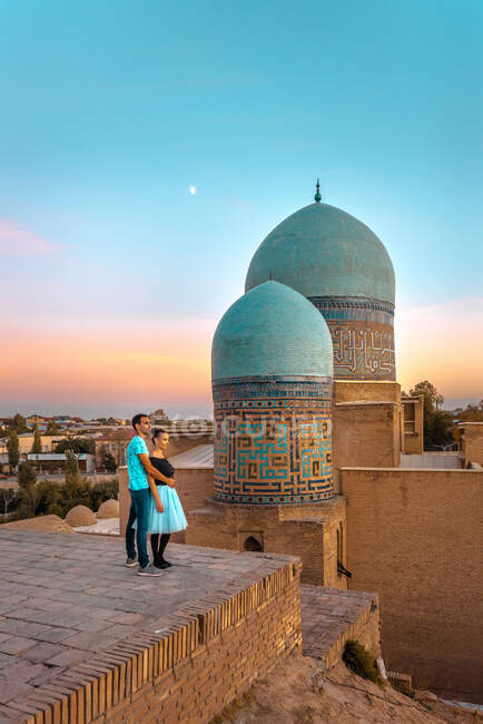 Пара, обнимающая друг друга возле древнего исламского здания с куполами во время посещения Шах-и-Зинды в Самарканде, Узбекистан — стоковое фото