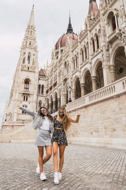 Mulheres alegres e elegantes passeando na praça de pedra com edifício antigo com torres pontiagudas e cúpulas em Budapeste — Fotografia de Stock