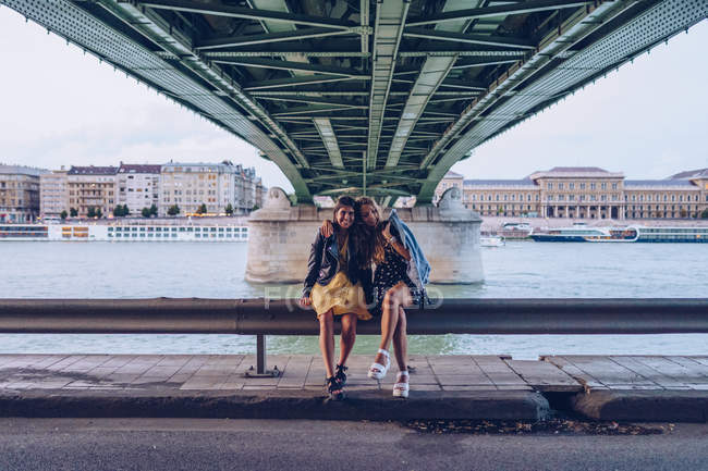 Активные веселые туристические девушки обнимаются, сидят на бетонном заборе под большим мостом и смотрят в камеру в Будапеште — стоковое фото