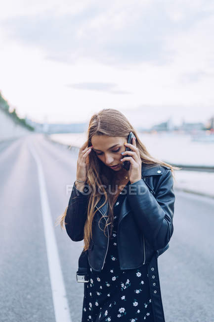 Femme sérieuse en veste noire redressant les cheveux et parlant sur téléphone portable sur la route vide à Budapest — Photo de stock
