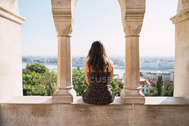 Vista trasera de la solitaria mujer de pelo largo sentada en una antigua cerca de mármol junto al jardín en la ciudad de Budapest - foto de stock