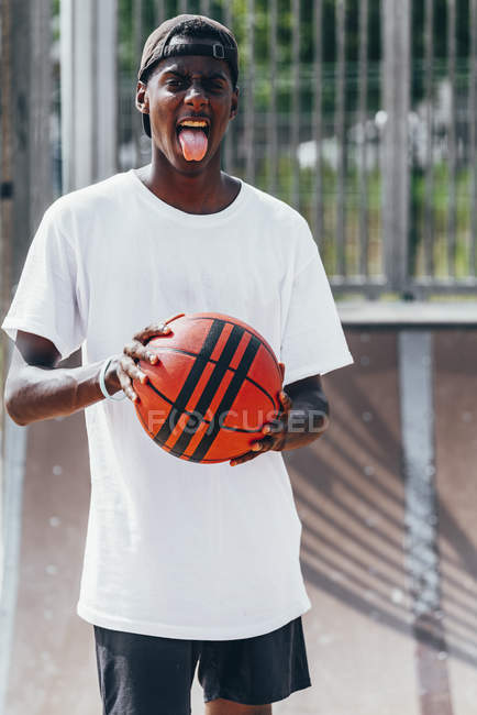 Веселый афроамериканец держит оранжевый мяч и смотрит в камеру с высунутым языком с широкой улыбкой — стоковое фото