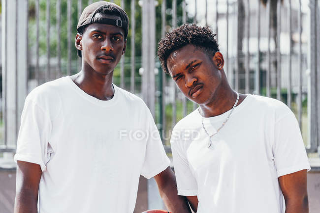 Serious chicos afroamericanos sosteniendo bola naranja y mirando a la cámara en día brillante cerca de valla de metal - foto de stock