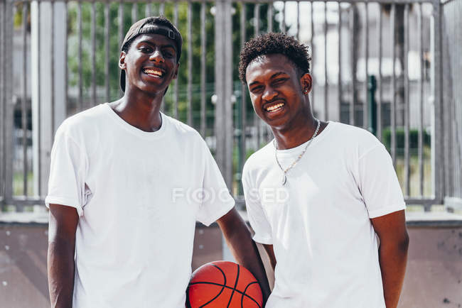 Happy African American guys holding orange ball and looking at camera in bright day near metal fence — Stock Photo