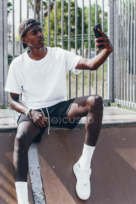 Pensivo sportivo afroamericano che si fa selfie con il telefono cellulare sulla recinzione del parco giochi nella giornata luminosa — Foto stock