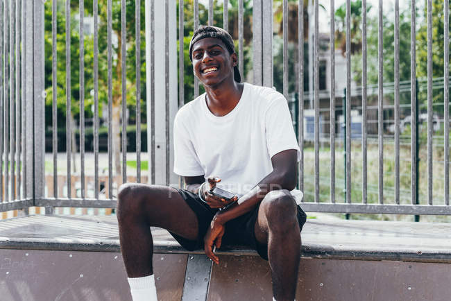 Afrikanischer amerikanischer Sportler surft bei hellem Tag mit Handy in Kamera am Zaun eines Spielplatzes — Stockfoto