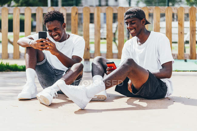 Divertidos e alegres esportistas afro-americanos surfando celular confortável colocado no playground em dia brilhante — Fotografia de Stock