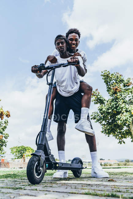 Africano americanos esportivos homens se divertindo piggybacking e andar em scooter elétrico em dia nublado brilhante — Fotografia de Stock