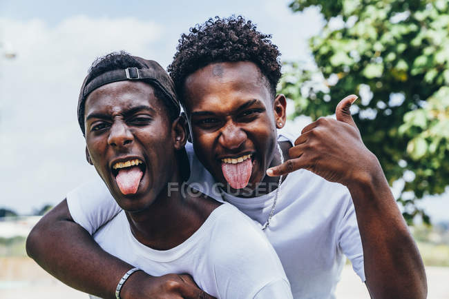Fröhliche Umarmung afrikanisch-amerikanischer junger Männer, die Zunge in die Kamera zeigen und mit Fratze im Gesicht gestikulieren — Stockfoto