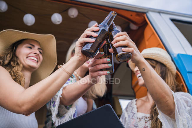 Улыбающиеся веселые дамы с длинными волосами в шляпах, поднимающие бутылки с напитками и звенящие на отдыхе в машине — стоковое фото