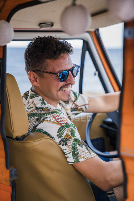 Stylish mustache man in sunglasses and colorful shirt holding steering wheel and smiling in camera in car on beach — Stock Photo