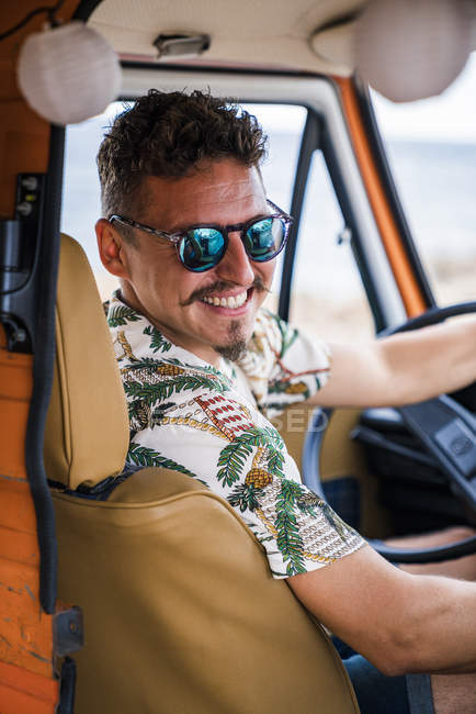 Elegante hombre bigote en gafas de sol y camisa colorida sosteniendo el volante y sonriendo en cámara en coche en la playa - foto de stock