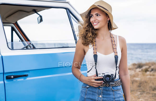 Encantadora senhora alegre em chapéu segurando câmera nas proximidades carro azul na praia e olhando ao redor — Fotografia de Stock