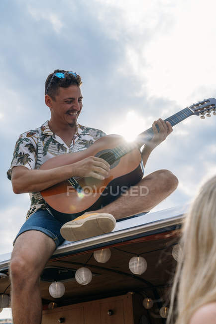 Homme enthousiaste jouant de la guitare assis sur le toit de la voiture tout en charmante dame profiter de la musique en voiture sur la plage — Photo de stock