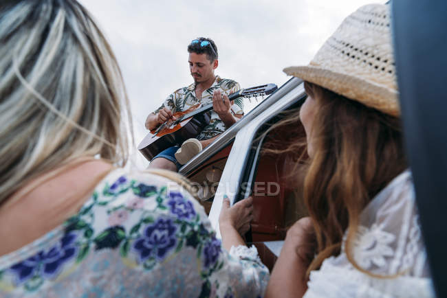 Hombre entusiasta tocando la guitarra sentado en el techo del coche mientras que las señoras encantadoras disfrutan de la música en coche en la playa - foto de stock