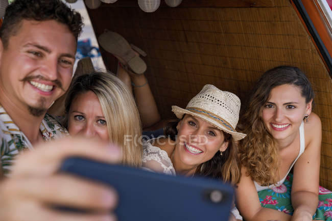 Pleasant group of young friends in trunk of minivan taking selfie on phone on beach in sunny daytime — Stock Photo