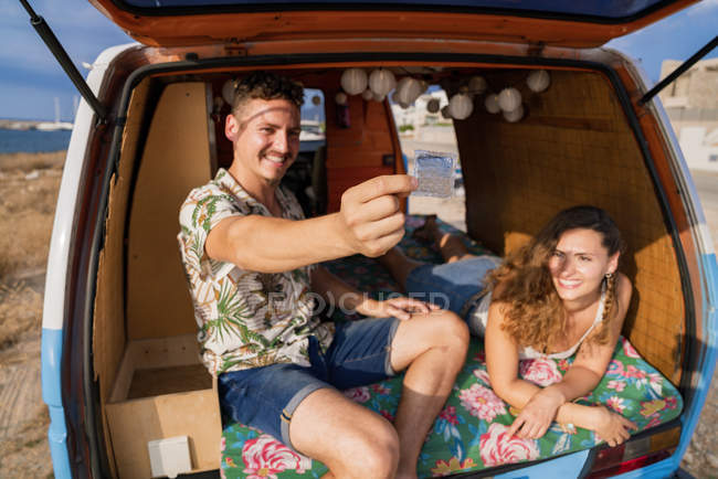 Радостная пара, показывающая презерватив и улыбающаяся в багажнике автомобиля — стоковое фото