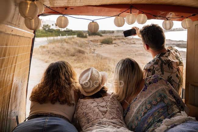 Agradable grupo de amigos en el maletero de minivan tomando selfie en el teléfono en la playa en el día soleado - foto de stock