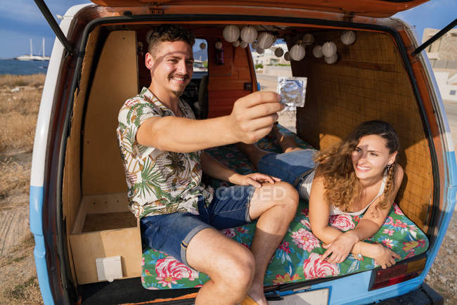 Alegre pareja cómoda colocación en el maletero del coche mostrando el condón y sonriendo - foto de stock