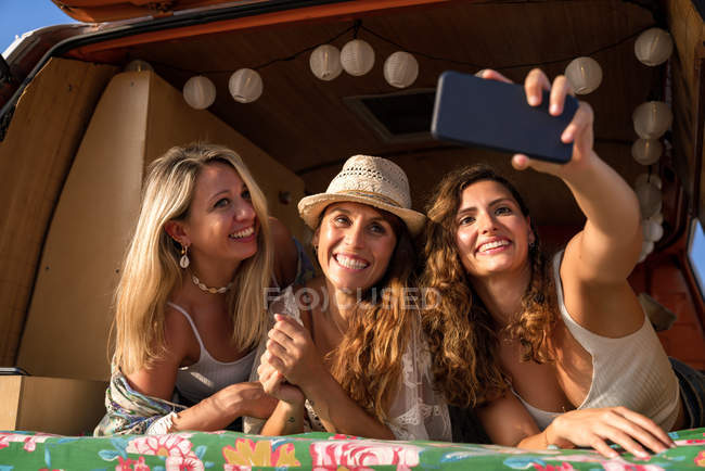 Le signore piacevoli allegre in tronco di minivan lucente che si divertono come prendere selfie su telefonino su spiaggia in giorno soleggiato — Foto stock