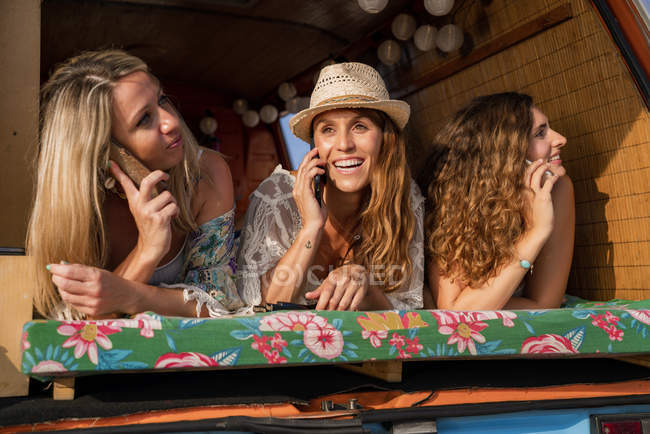 Усміхнені приємні дами лежать на багажнику яскравого мінівена і розмовляють на мобільних телефонах на пляжі в сонячний день — стокове фото