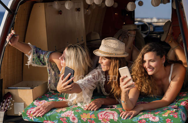 Joyeux dames agréables couché sur le tronc de minifourgonnette lumineuse et avoir du plaisir à prendre des selfies sur les téléphones mobiles sur la plage — Photo de stock