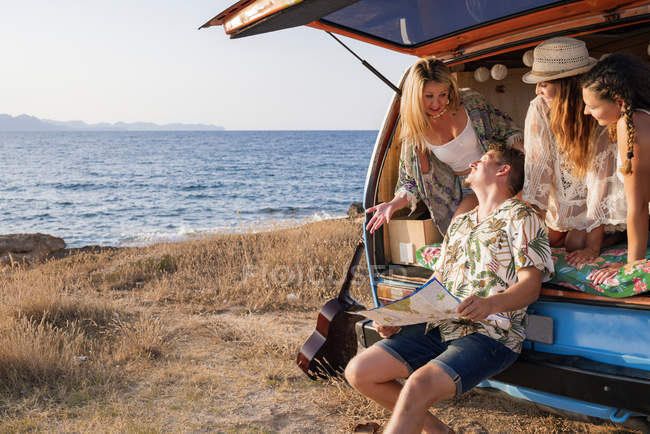 Curios activos hombre mirando hoja de ruta abierta y encontrar ruta con mujeres encantadoras en sombreros en tronco abierto de coche en la playa - foto de stock