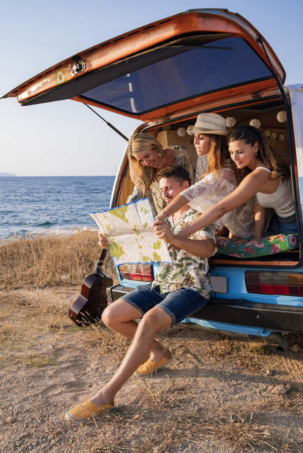 Active curios man looking at open road map and finding route with charming women in hats in open trunk of car on seaside — Stock Photo