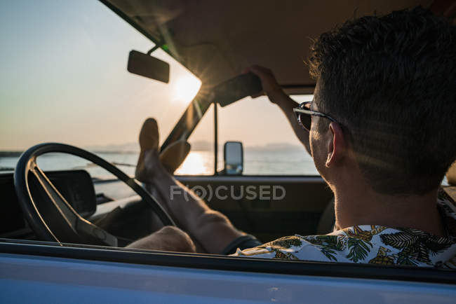 Cheerful male in sunglasses taking selfie with smartphone while sitting relaxed with legs on dashboard in parked car — Stock Photo