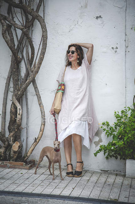 Optimistic woman with raised arm leaning on wall in Marbella beside beautiful tree and holding greyhound on leash — Stock Photo