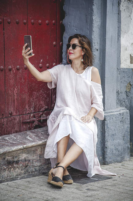 Confident stylish woman in sunglasses and modern summer dress taking selfie with mobile phone while sitting on stone door sill — Stock Photo