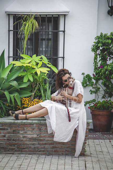 Vue latérale d'une femme calme câlinant un chien tout en se reposant ensemble sur une clôture de parterre de fleurs avec des plantes tropicales à Marbella — Photo de stock